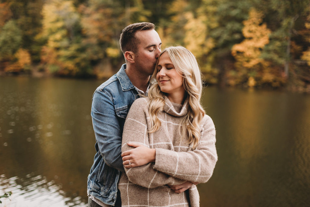 man kisses woman on top of head in front of fall colors and lake during their brown county engagement session