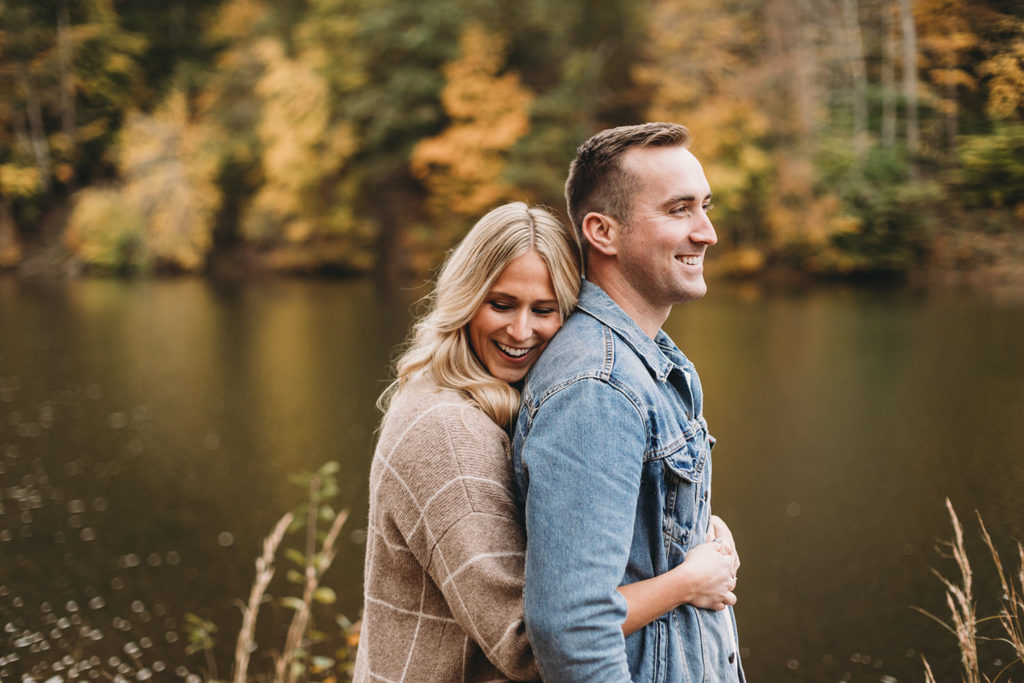 woman hugs man from behind as they smile in front of leaves that have changed color for fall during their brown county engagement session