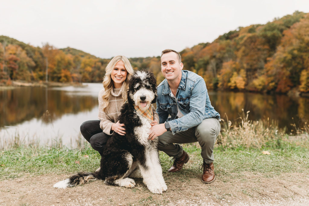 man and woman crouch and pet dog in front of ogle lake which is reflecting the colors of the leaves in fall during their brown county engagement session