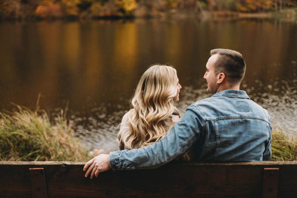 man with arm around woman sitting on bench in front of ogle lake in fall during their brown county engagement session