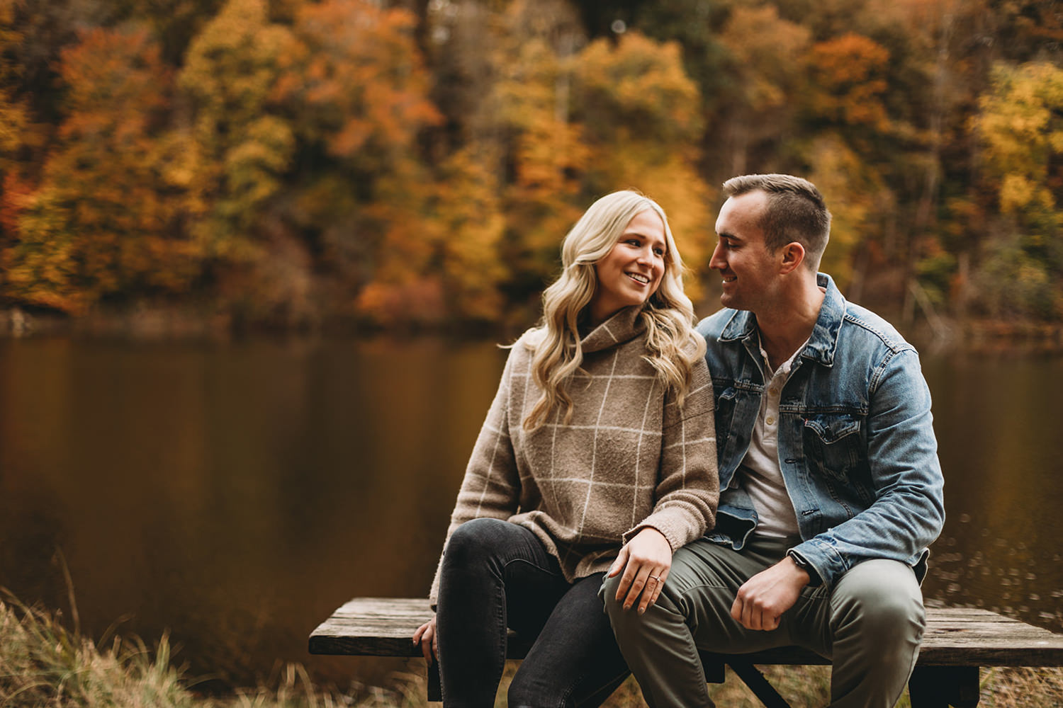 man in jean jacket and woman in sweater sit on picnic bench in front of lake and lots of fall foliage during their brown county engagement session