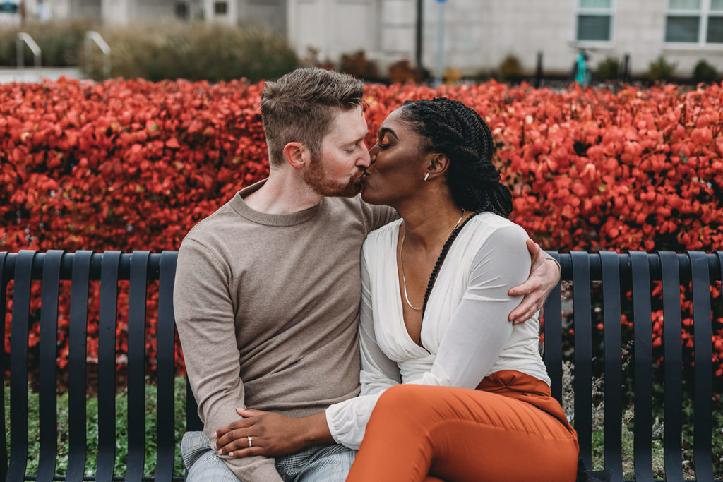 a man and a woman sit on a park bench and kiss in front of red bushes during their Carmel City Center Photography session