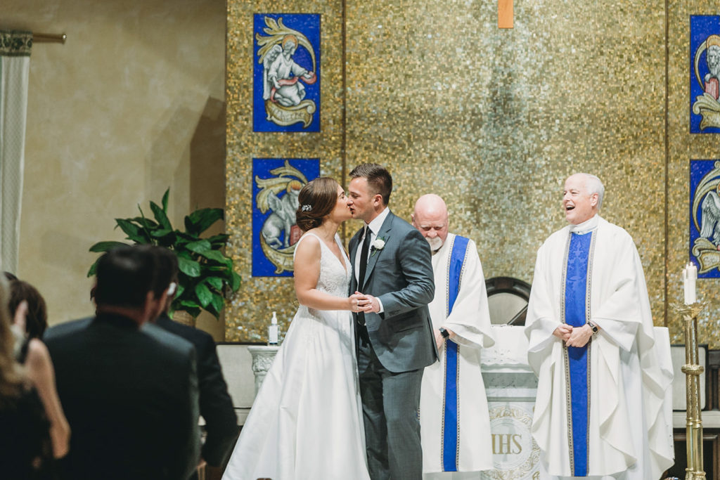 bride and groom share first kiss while priests watch during their charming carmel wedding