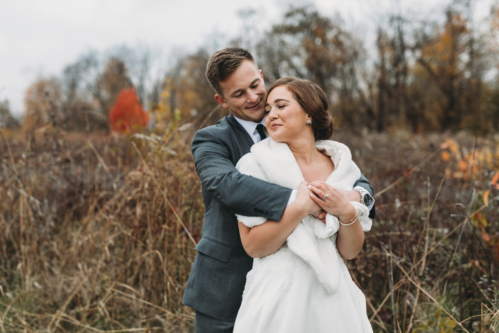 man in gray suit hugs woman in white dress with faux fur shawl from behind in meadow in autumn during their charming carmel wedding