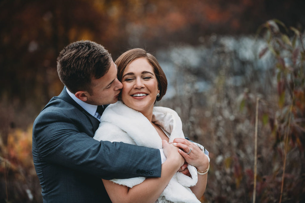 red leaves provide splashes of color in the background as a groom kisses his bride on the cheek while hugging her from behind on a crisp autumn day during their charming carmel wedding
