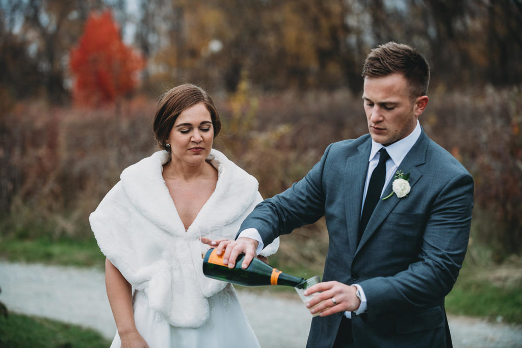 groom pours champagne in glass in meadow during their charming carmel wedding