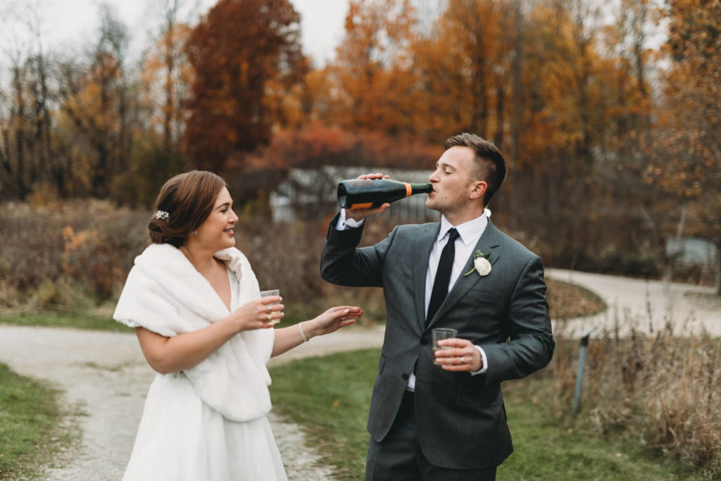 bride and groom drink champagne from glasses and from bottle in a meadow in cool creek park in fall with lots of bright fall colors behind them during their charming carmel wedding