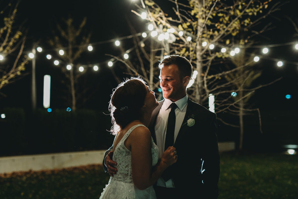 bride and groom hug and groom smiles at bride whilst backlit by string lights hanging off tree during their charming carmel wedding