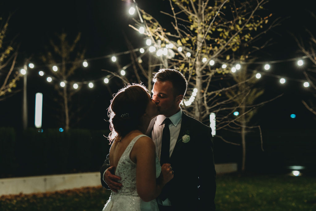 bride and groom kiss in front of tree lit by string lights at night during their charming carmel wedding