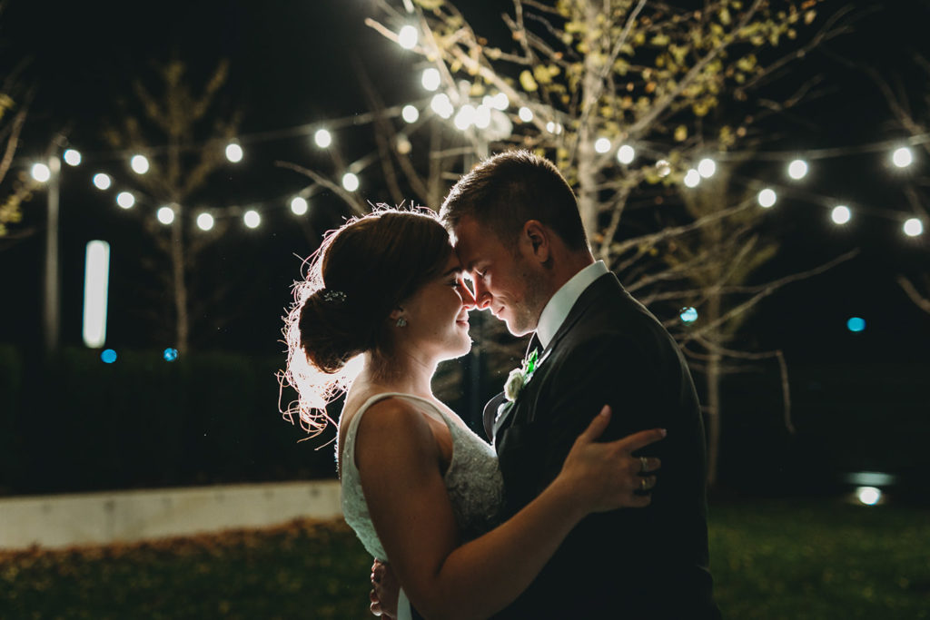 bride and groom touch foreheads and are backlit by string lights during their charming carmel wedding