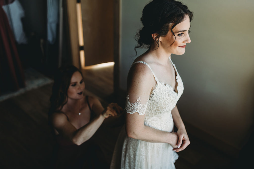 bridesmaid helps bride button up her dress in a dark and moody getting ready photo during their charming Lindley Farmstead wedding