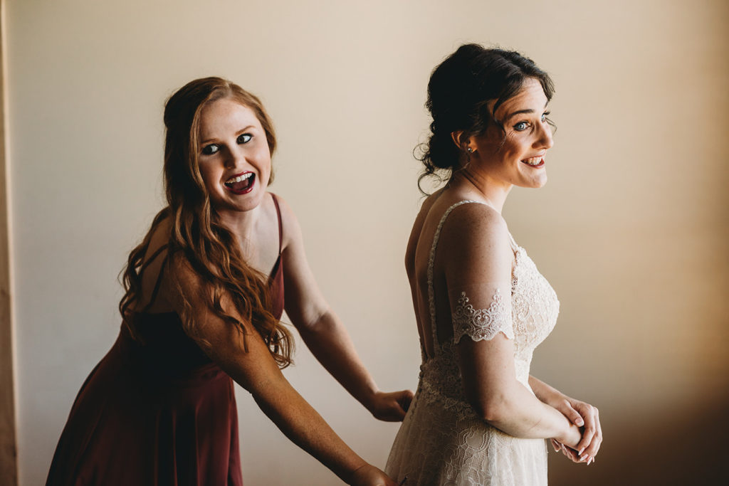 bridesmaid smiles and laughs as she helps bride into dress in window light during their charming Lindley Farmstead wedding