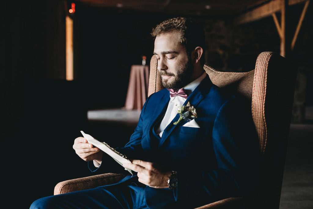 groom sitting in cushioned chair in window light reading letter from bride during their charming Lindley Farmstead wedding