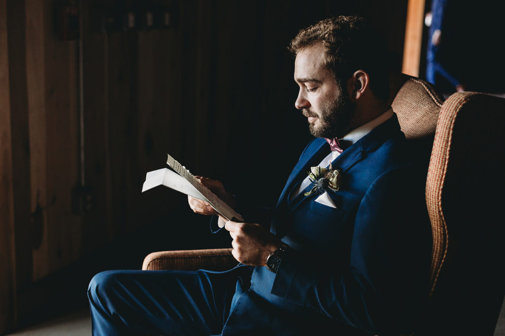 groom in blue suit sitting in chair in window light in basement during their charming Lindley Farmstead wedding
