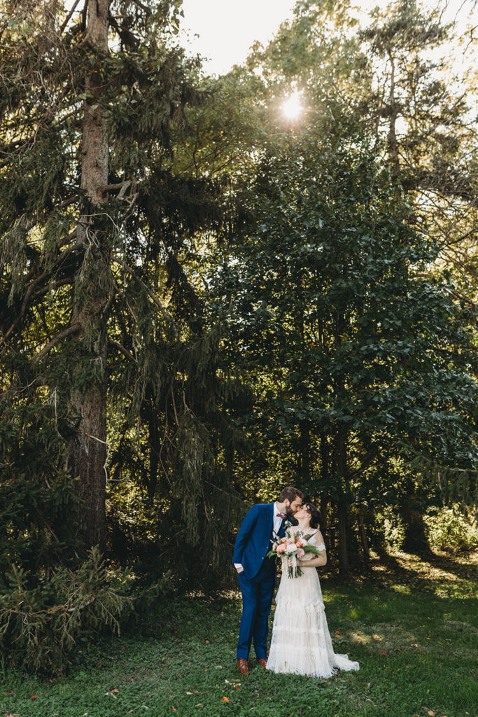 sun begins its descent behind trees as bride and groom kiss during their charming Lindley Farmstead wedding