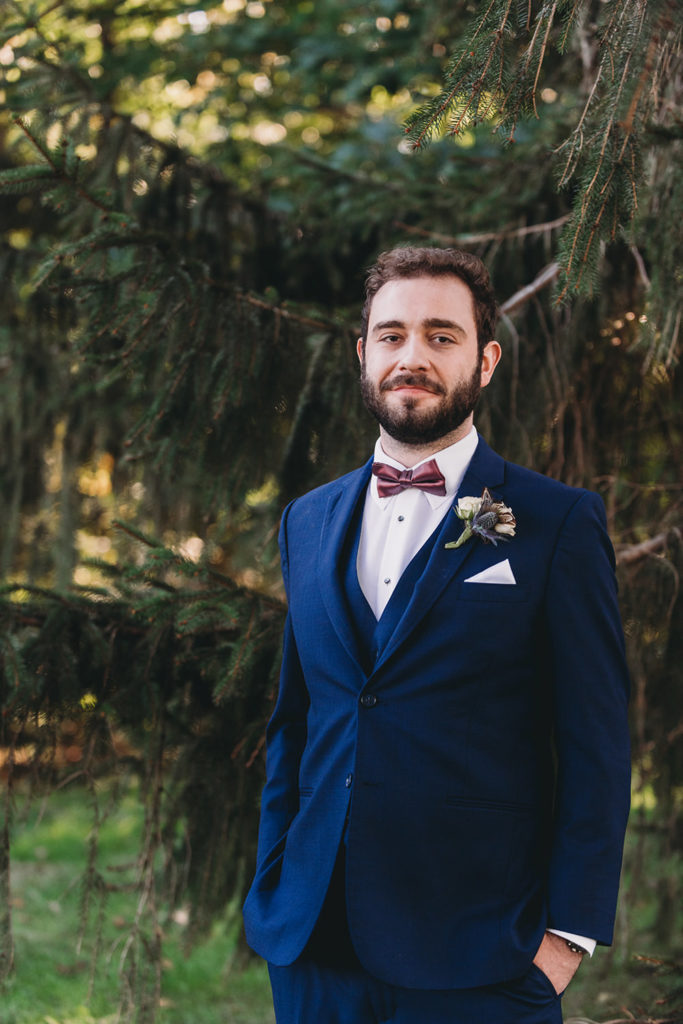 handsome groom in blue suit with burgundy bow tie in front of evergreen trees during their charming Lindley Farmstead wedding