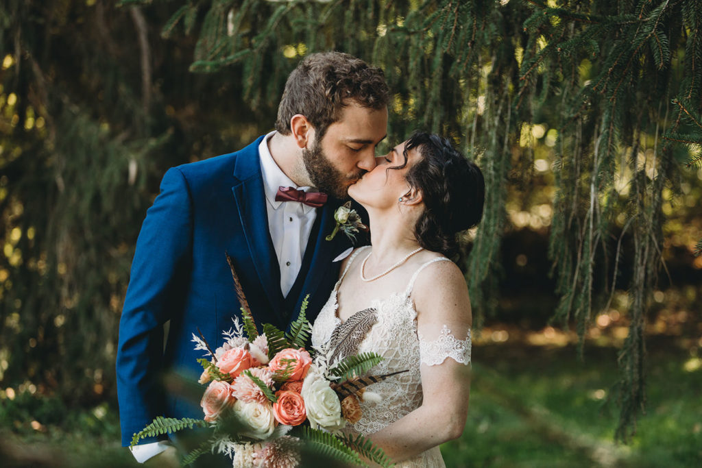 bride and groom kissing in front of evergreen trees during their charming Lindley Farmstead wedding