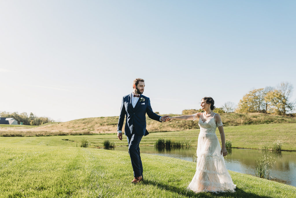 groom leads bride by the hand away from a pond during their charming Lindley Farmstead wedding