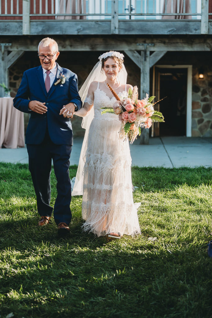 father of bride walks bride in bohemian dress with flower crown down the aisle during their charming Lindley Farmstead wedding
