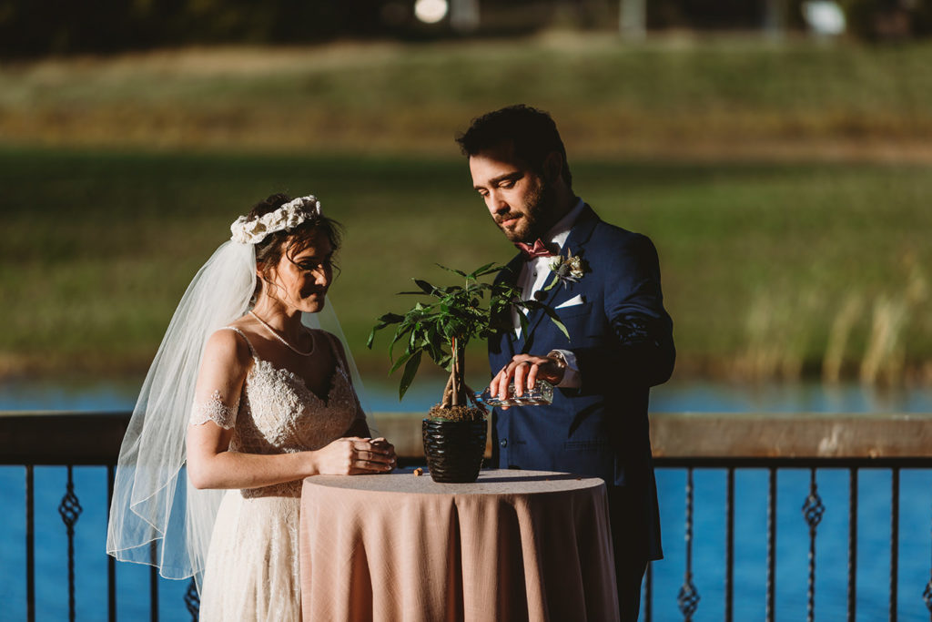 bride in bohemian dress and groom in blue suit water a plant together in this dark and moody ceremony photo during their charming Lindley Farmstead wedding