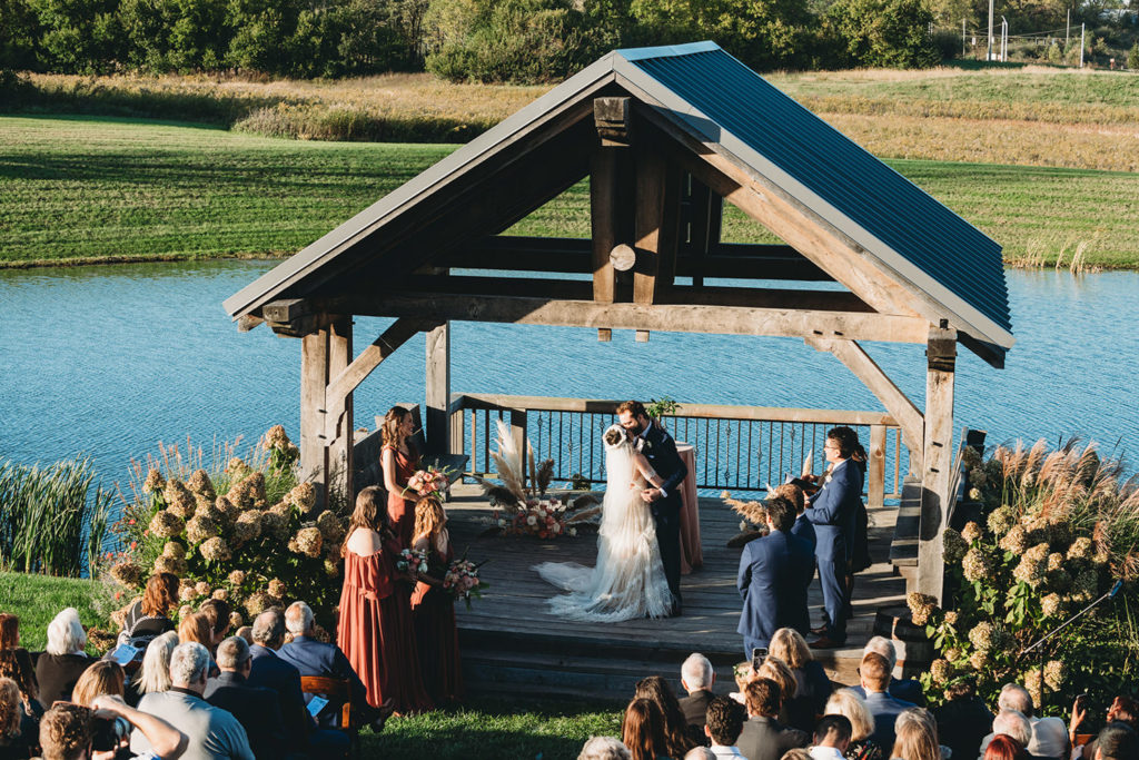 bride and groom share a first kiss under a gazebo in front of a pond during the wedding ceremony while guests watch during their charming Lindley Farmstead wedding