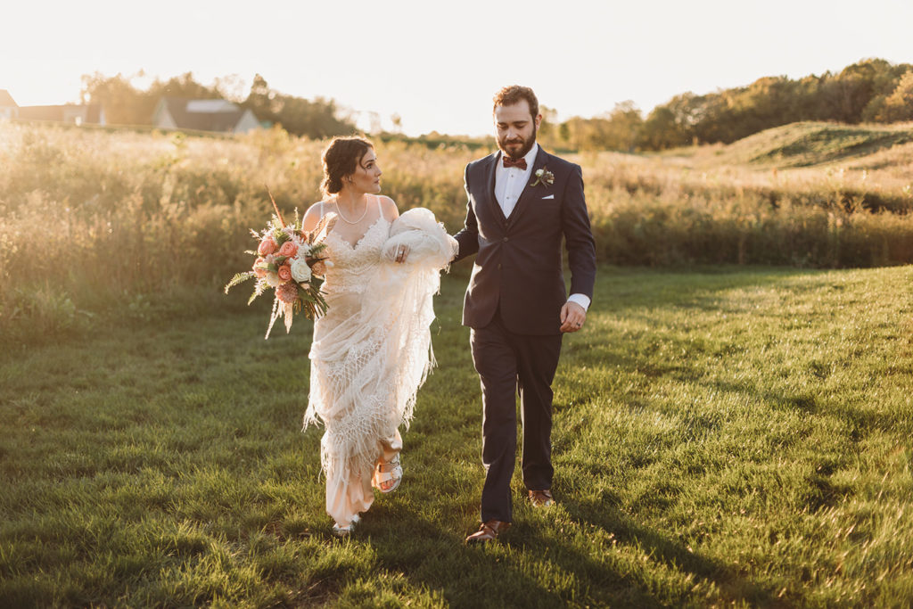 bride and groom walk through grass in front of a golden field at sunset during their charming Lindley Farmstead wedding