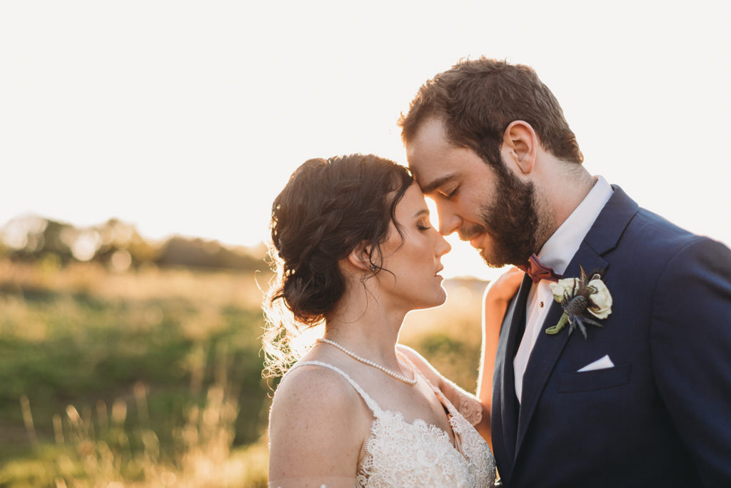 bride and groom touch noses with eyes closed in front of a golden field at sunset during their charming Lindley Farmstead wedding