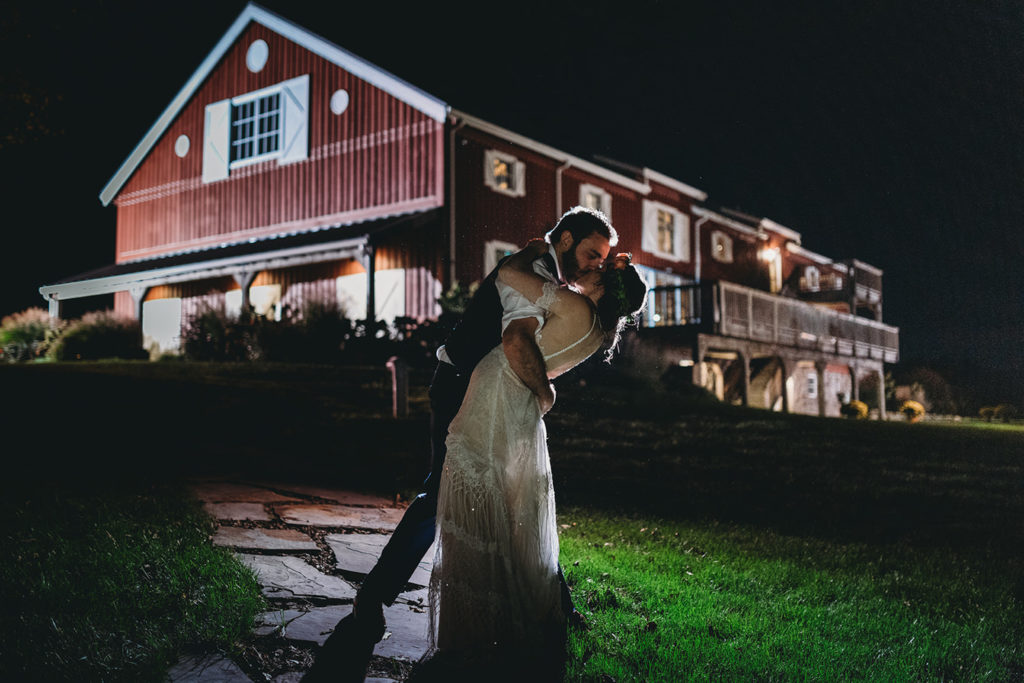 bride and groom kiss in front of red barn at night during their charming lindley farmstead wedding, one of the Best Indianapolis Barn Venues