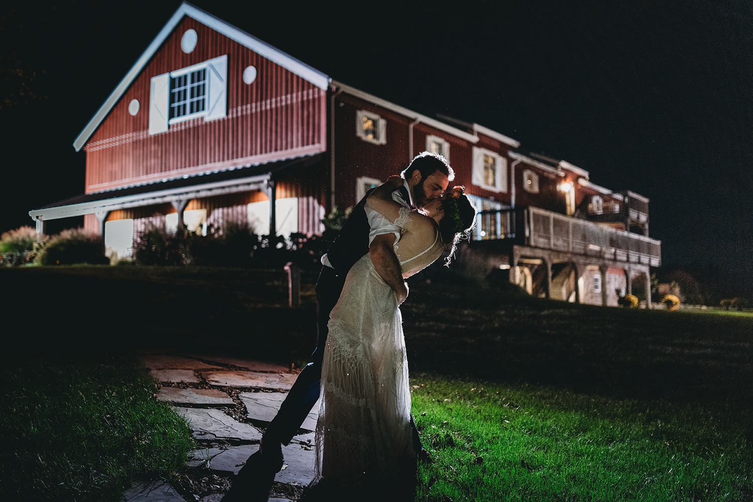 bride and groom kiss in front of red barn at night during their charming lindley farmstead wedding