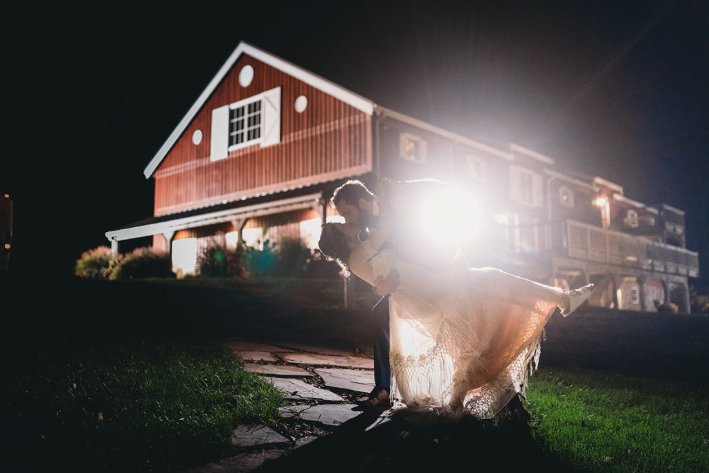 groom dips and kisses bride as flash pops off behind him lighting up a giant red barn during their charming Lindley Farmstead wedding