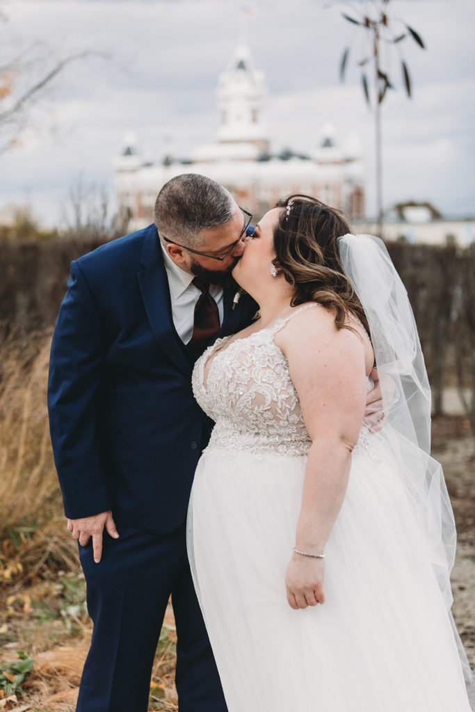 bride and groom kiss while clock of court house is visible behind them in franklin indiana during their garment factory events wedding