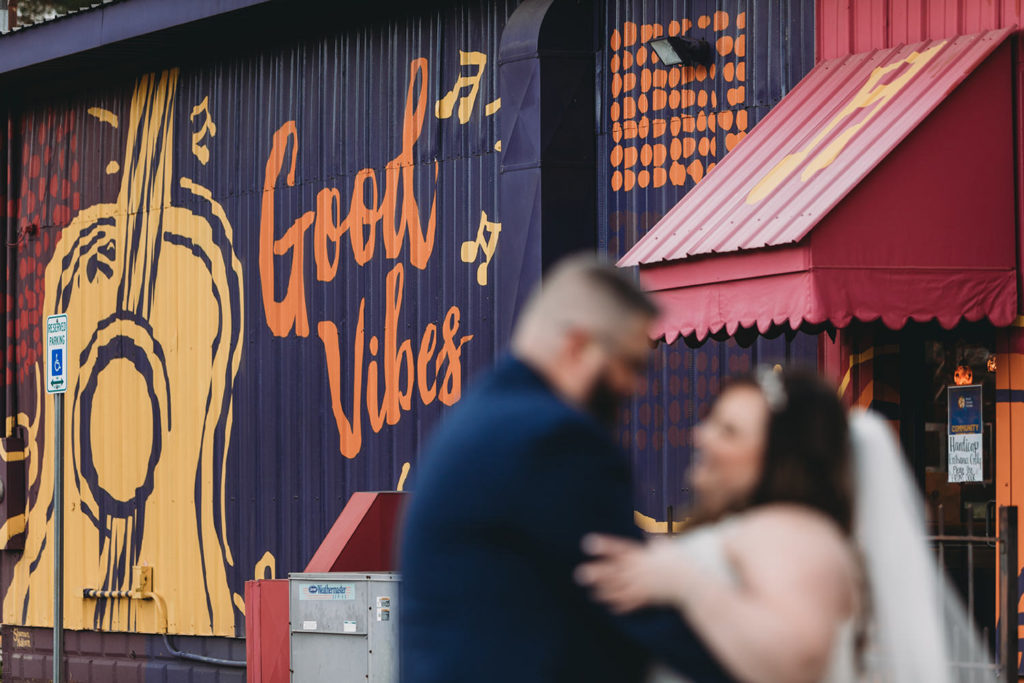 sid eof building says good vibes and has giant yellow guitar painted on it and bride and groom are out of focus in front of it during their garment factory events wedding