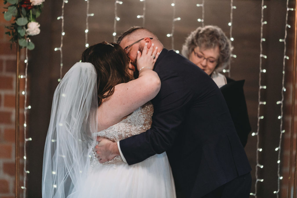 bride and groom first kiss in front of string lights and copper arch with floral during their garment factory events wedding
