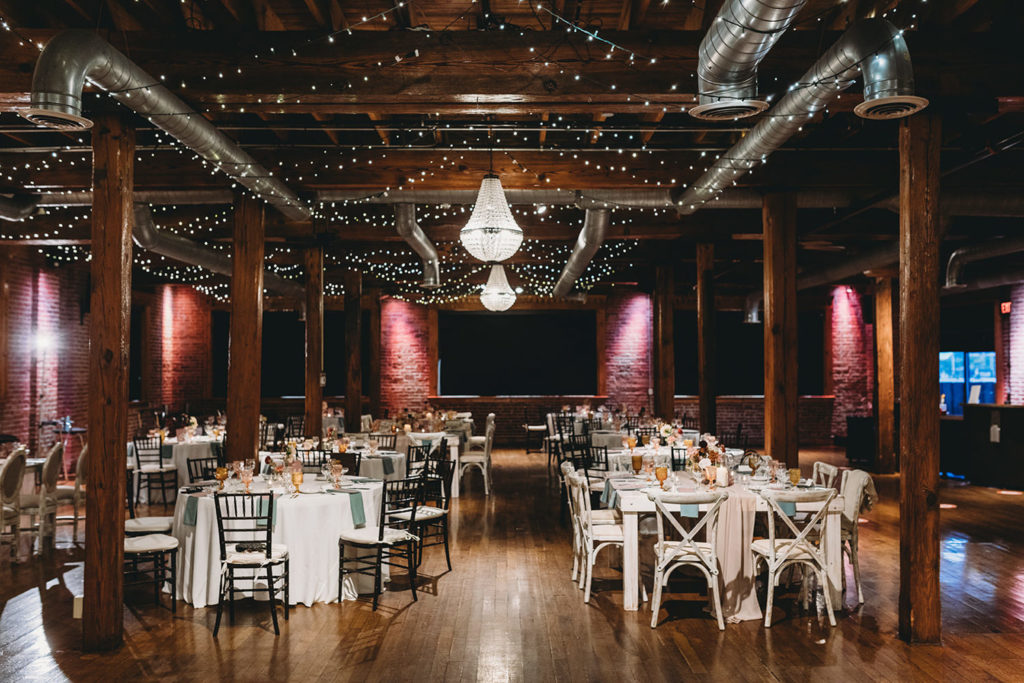 pink mood lighting on brick walls and a flash or two light up the mavris event space covered in string lights and chandeliers and tables and chairs during their mavris reception