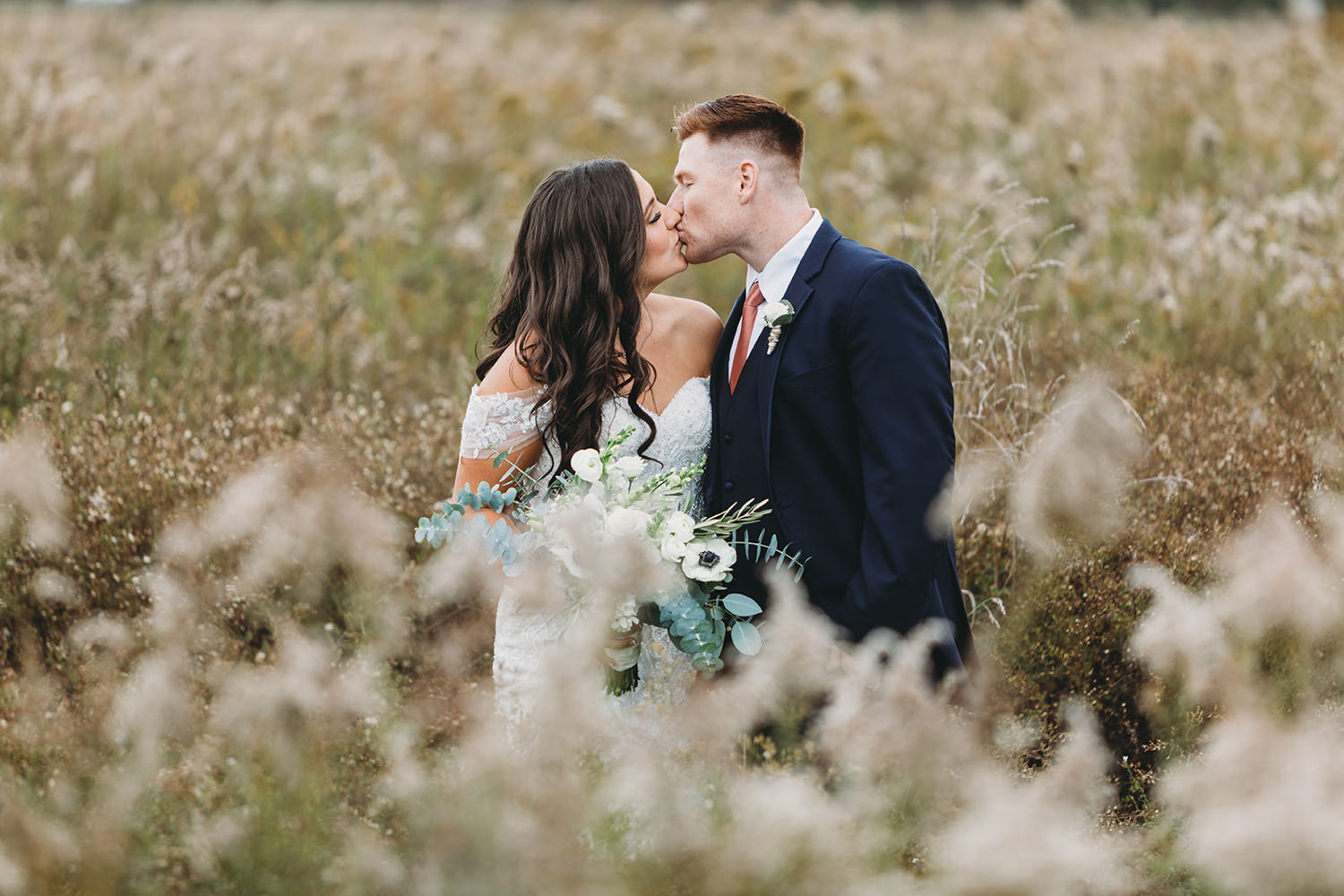 man in suit and woman in white dress with lace kiss in the middle of a field of wildflowers during this White Willow Farms Wedding