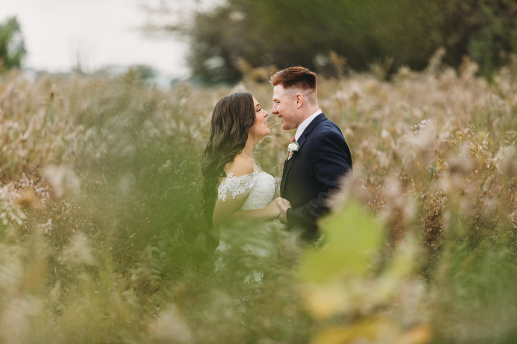 green flora in the foreground is out of focus and frames a bride and groom in a field of brown wildflowers during their white willow farms wedding