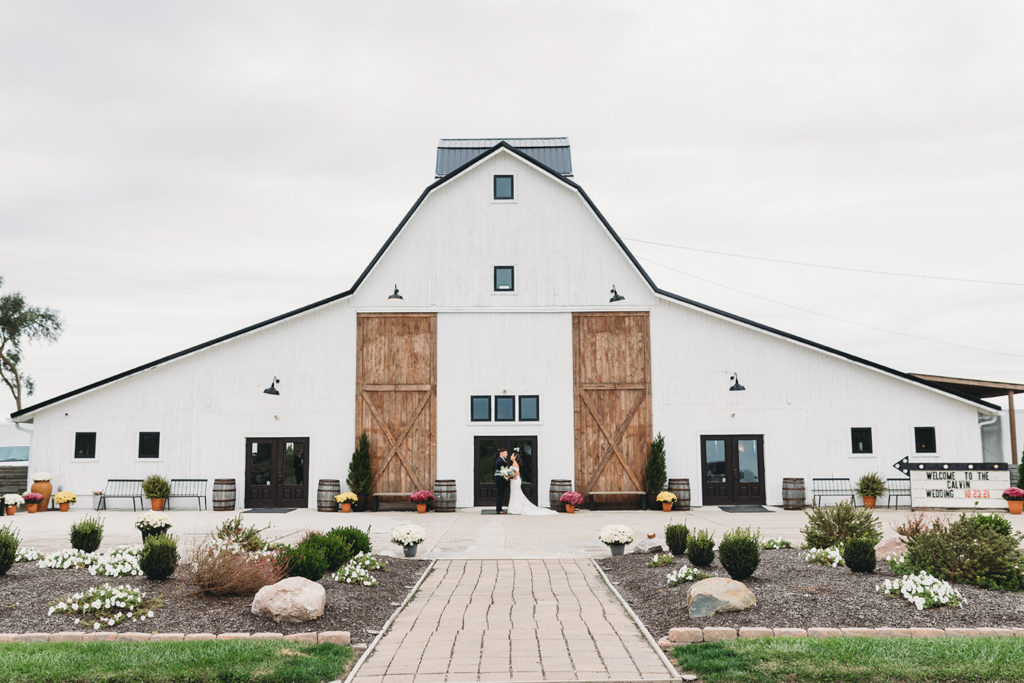 wide shot of bride and groom kissing in front of a large white barn during their white willow farms wedding