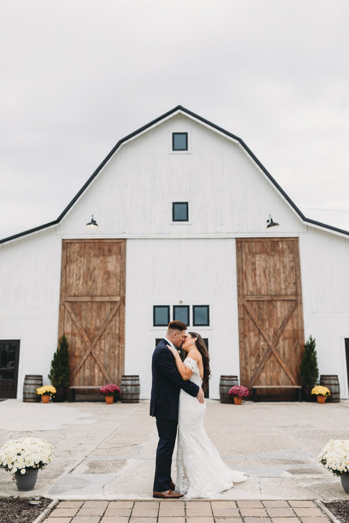 vertical oriented photo of bride and groom kissing in front of white barn with large brown wooden doors during their white willow farms wedding