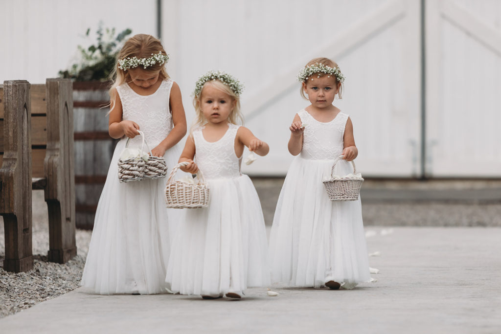 three blonde flower girls who are very young walk down an aisle dropping flowers during their white willow farms wedding