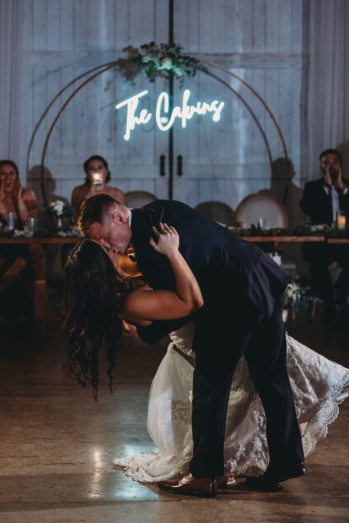 groom dips and kisses bride in front of wooden circle standing upright with their name hanging down in neon during their white willow farms wedding