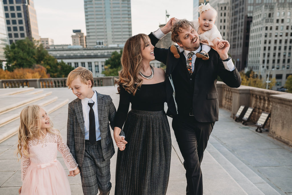 From left to right: A little girl, a preteen boy, a mom, a dad, and a baby on dad's shoulders walk hand in hand with the downtown Indianapolis city in the background. the mom and dad are multitalented wedding photographers