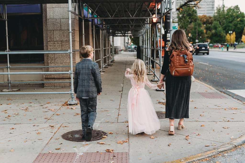 A mom with a backpack holds her daughters hand along a busy city street while her son walks on the other side of the daughter. They all wear formal wear. a female multitalented wedding photographers carrying a photography bag with her kids