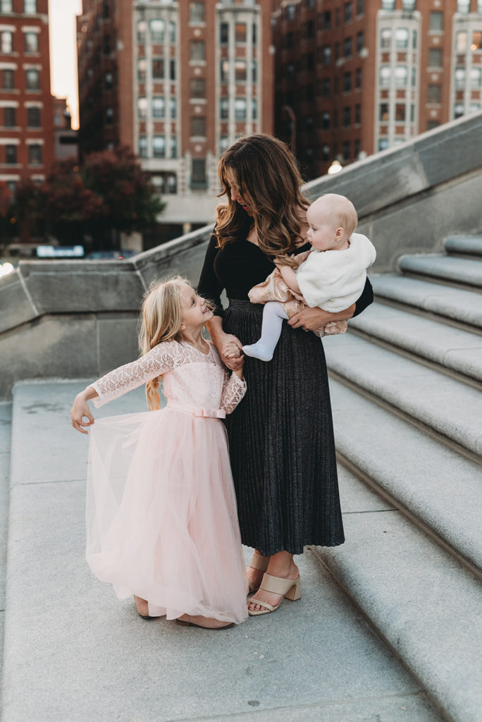 mom stands with and looks down and smiles at daughter who is holding her dress out, showing it off. The mom hold a baby on her other hip. They are standing on a set of outdoor stairs with buildings in the background.