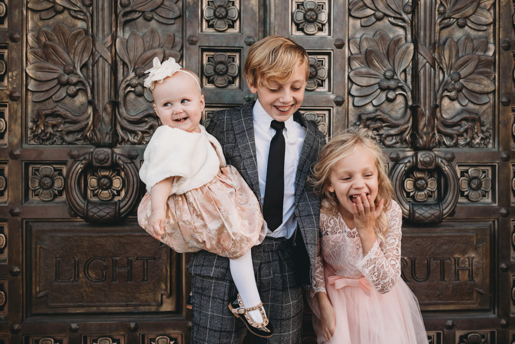 A baby, pre-teen boy, and little girl stand in front of a gold door while they giggle. The little girl has her hands over her mouth.