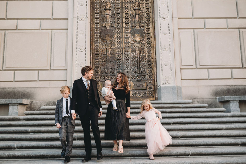 A family of five walk down a set of stone staircases. There is a large gold door behind them with stone walls. Son and dad are in suits and the mom and daughters are in dresses. Check out these multitalented wedding photographers