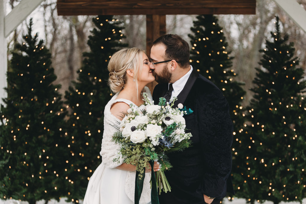 Bride and groom kissing in front of lit christmas trees and a wooden cross.
