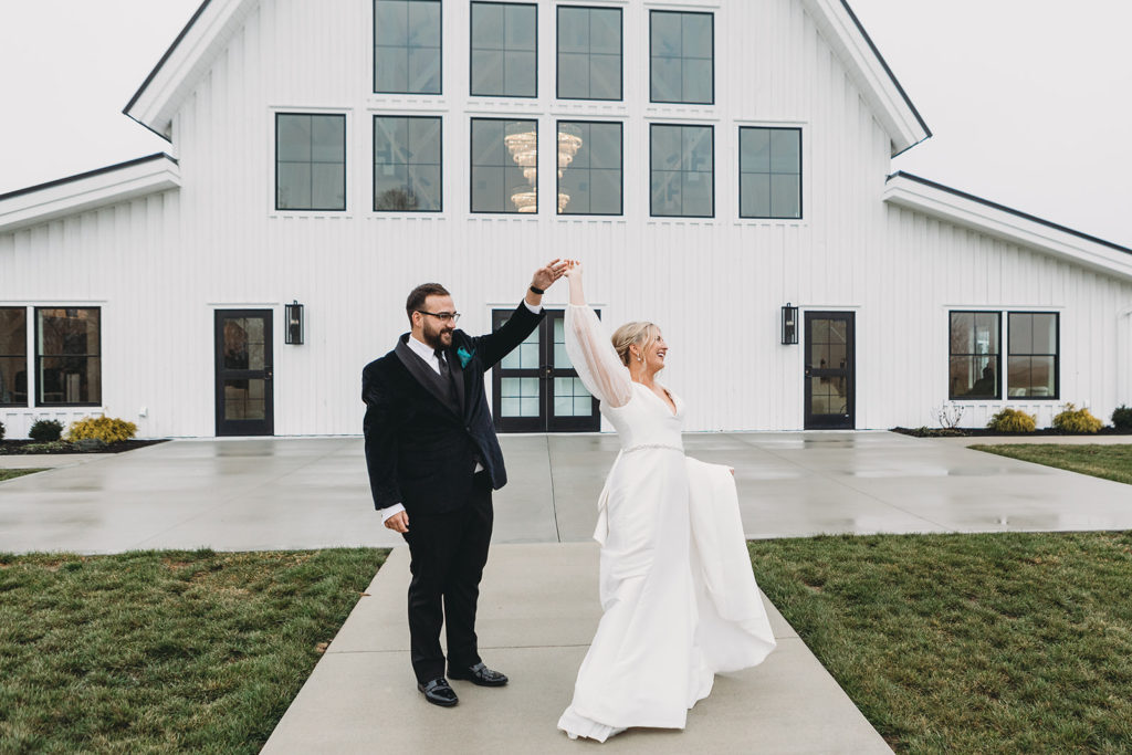 The groom twirls his wife in front of the white sixpence venue in Indianapolis between two strips of green grass at their wedding.
