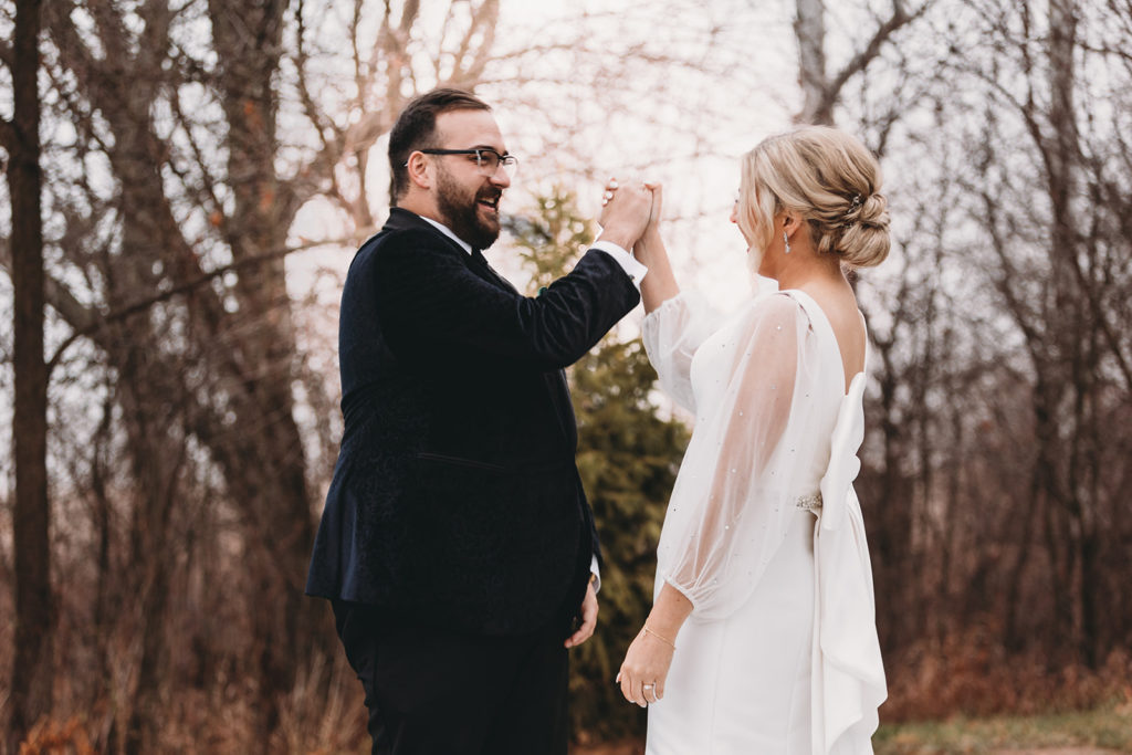 Bride and groom give each other a high five while facing one another in front of a tree line while the sun sets.