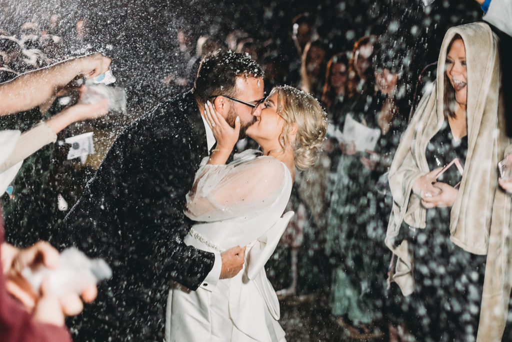 Bride and groom kiss while people throw snow on them outdoors.