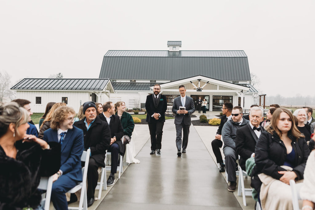 Groom making his way down the outdoors aisle with the officiant while the guest look on each side. Pavement is shiny from the rain moments before the ceremony.